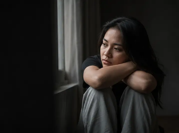 Wanita muda duduk merenung di dekat jendela dalam suasana redup yang menggambarkan proses hipnoterapi untuk pemulihan trauma